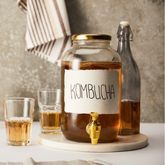 Bouteille et bocal de kombucha posés sur une table avec deux verres remplis de boisson ambrée et un morceau de gingembre en premier plan.