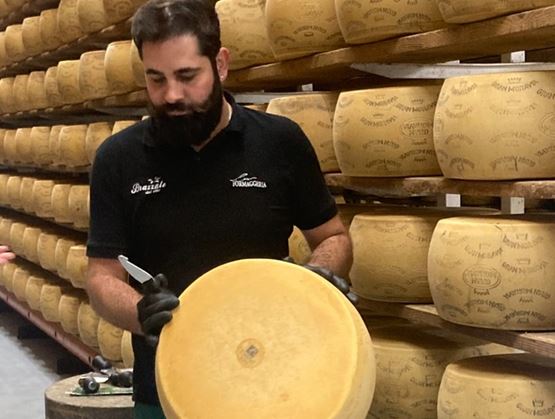Fromager dans une cave d’affinage manipulant une grande meule de fromage, entouré de rangées de fromages affinés sur des étagères en bois.