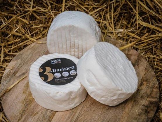 Trois fromages à croûte blanche posés sur une planche en bois, sur fond de paille, dont un avec une étiquette « Le Parisien » de la fromagerie Dongé.