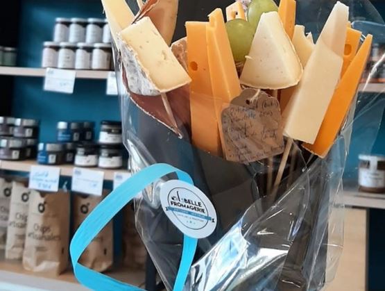 Bouquet gourmand composé de morceaux de fromage et de raisins présentés sur des piques, emballé dans un film transparent avec un ruban bleu, devant des étagères d’épicerie fine.