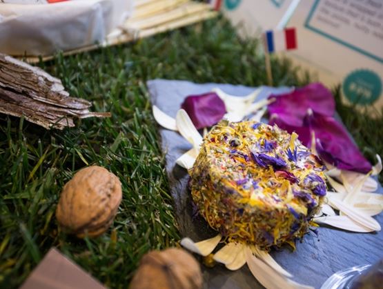 Fromage rond présenté sur une ardoise, enrobé de fleurs séchées colorées, au milieu d’une mise en scène végétale avec herbe, pétales, noix et petit drapeau français.