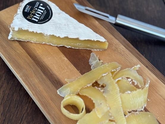 Part de fromage « Brie noir » posée sur une planche en bois, avec des copeaux de fromage au premier plan et un couteau à lame fine à côté.