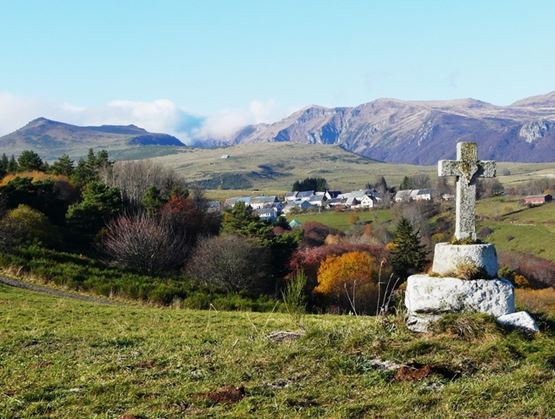 Paysage de montagne avec un village au loin et une croix en pierre au premier plan.