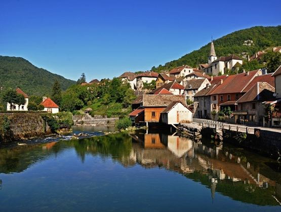 Village pittoresque au bord d’une rivière avec maisons et église entourées de collines verdoyantes.