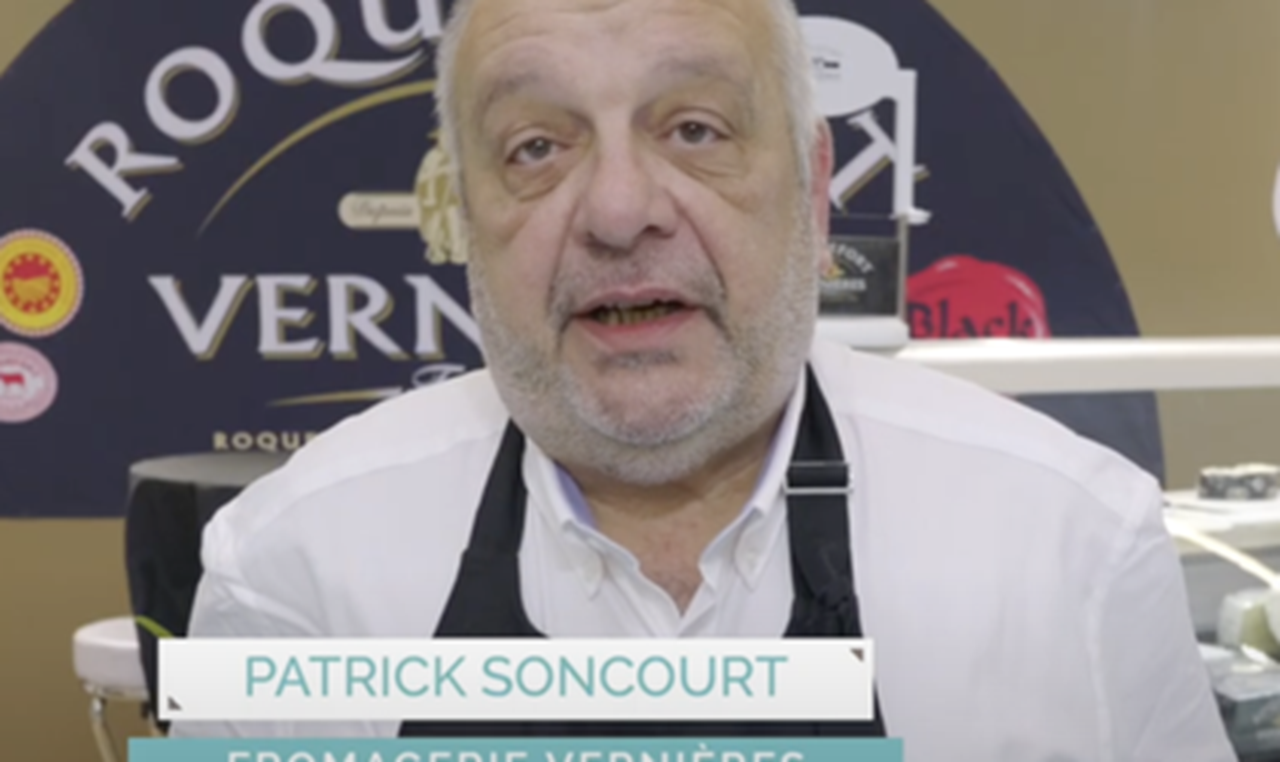 Fromager en tablier présentant son activité devant un stand de fromages.