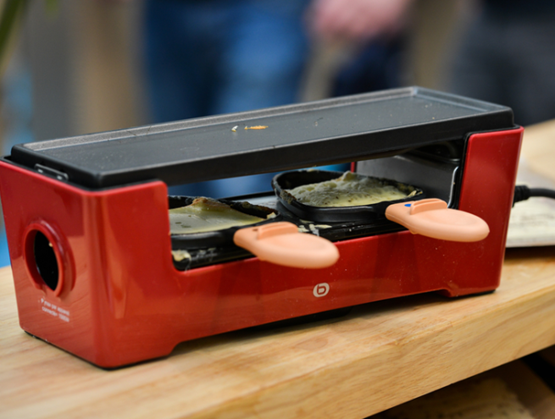 Mini appareil à raclette rouge avec poêlons individuels en cours de chauffe sur un plan de travail.