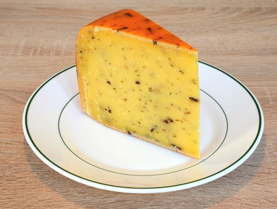 Part de fromage à pâte pressée à croûte orange, présentée sur une assiette blanche, avec des éclats d’herbes ou d’épices dans la pâte.