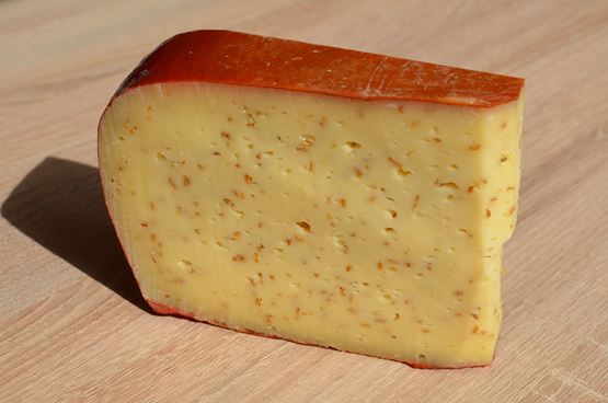 Morceau de fromage à pâte pressée à croûte orange, présenté sur une table en bois, avec de petits éclats répartis dans la pâte.
