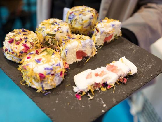 Petits fromages frais décorés de fleurs et d’herbes séchées, présentés sur une ardoise avec un cœur de fruit visible dans certaines portions.