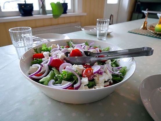 Salade grecque dans un grand bol avec laitue, tomates cerises, oignons rouges et cubes de feta, posée sur une table avec des verres et des assiettes.