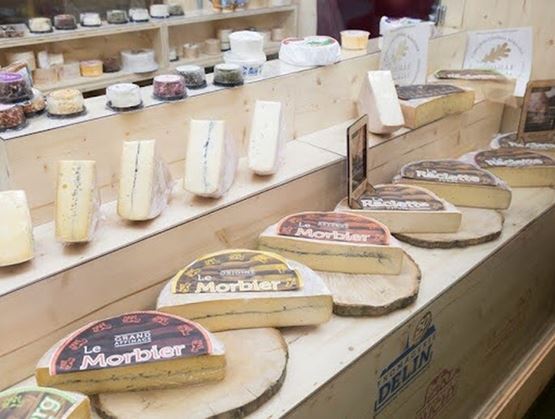 Un étal de fromages dans une fromagerie présentant différentes meules et morceaux de fromages, dont du Morbier, disposés sur des planches en bois avec des produits variés en arrière-plan.