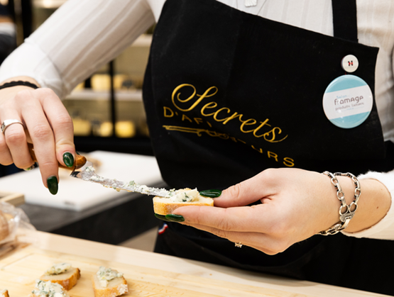 Une personne tartine du fromage sur des morceaux de pain à l’aide d’un couteau, portant un tablier noir dans un espace de dégustation ou de vente de fromages.