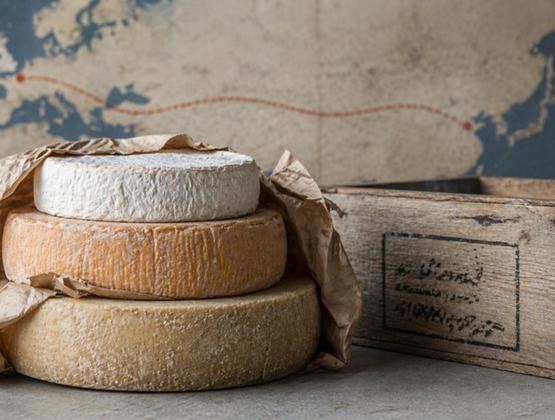 Trois meules de fromage empilées, dont un fromage à croûte fleurie et deux fromages affinés, posées sur une surface rustique avec du papier d’emballage et une caisse en bois, sur fond de carte ancienne.