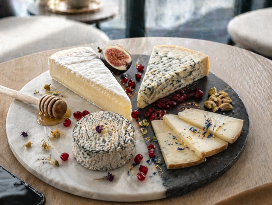 Plateau de fromages gourmets présenté sur une assiette en marbre, avec différents fromages (brie, fromage bleu, fromage affiné et fromage de chèvre), accompagné de miel, de figues, de graines de grenade, de pistaches et de fleurs comestibles sur une table en bois.