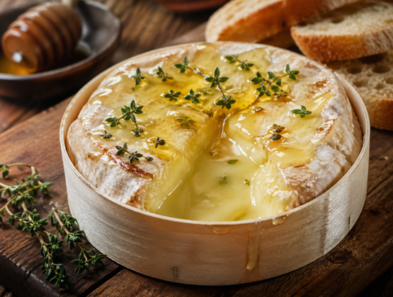 Fromage fondu dans sa boîte en bois, garni de miel et de thym, accompagné de tranches de pain sur une table en bois.