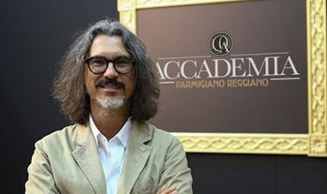 Homme posant les bras croisés devant un panneau Accademia Parmigiano Reggiano lors d’un salon professionnel.