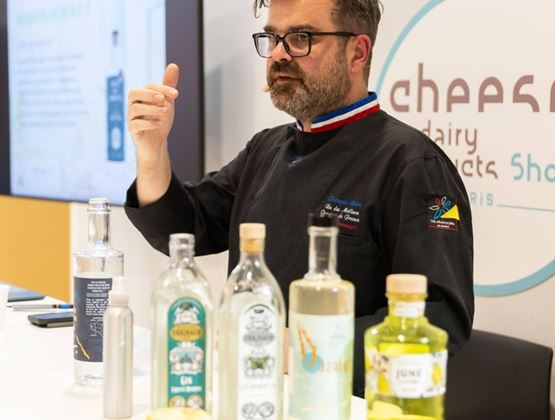 Expert fromager animant une présentation au Salon du fromage et des produits laitiers à Paris, avec des bouteilles et des fromages exposés sur la table.