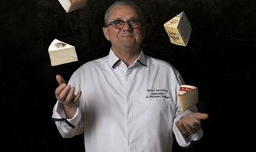 Maître fromager en veste blanche présentant plusieurs morceaux de fromage sur fond noir.