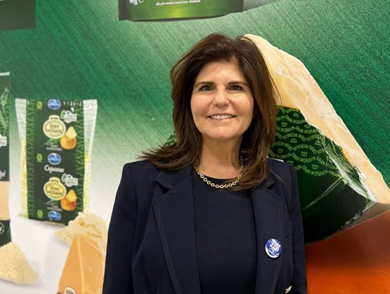 Portrait d’une femme souriante devant un stand présentant des fromages et une grande meule de fromage.