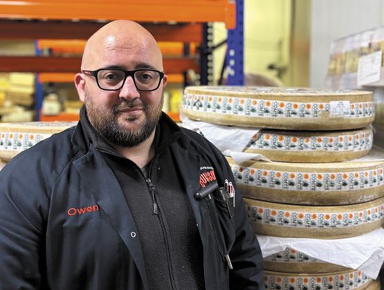 Portrait d’un professionnel en entrepôt, debout devant des meules de fromage empilées et emballées.
