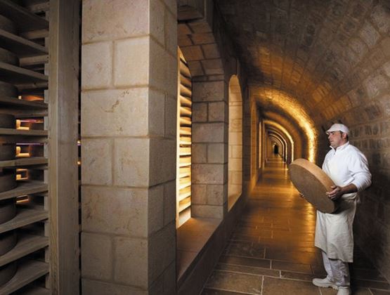 romager portant une meule de fromage dans une cave d’affinage voûtée, avec des fromages rangés sur des étagères.
