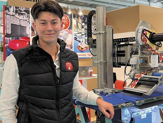 Portrait d’un jeune homme souriant sur un stand professionnel, à côté d’une machine d’étiquetage.