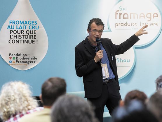 Un homme présente une conférence avec un micro devant un public, sur un stand consacré aux fromages au lait cru et aux produits laitiers.