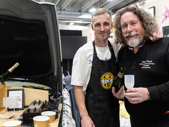 Deux hommes souriants posent dans un espace d’animation avec des bouteilles et du matériel de sonorisation, lors d’un événement professionnel.