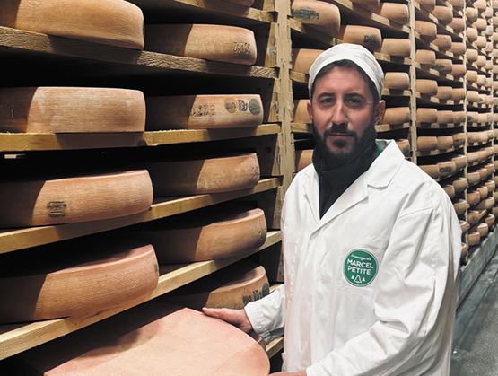 Fromager en tenue blanche posant dans une cave d’affinage, entouré de grandes meules de fromage rangées sur des étagères en bois.