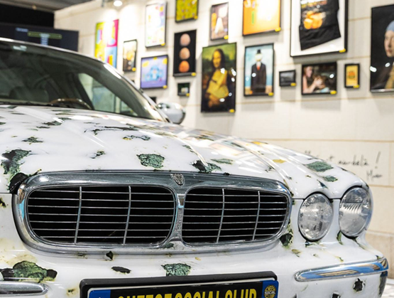 Voiture décorée sur le thème du fromage, exposée avec une plaque “Cheese Social Club” devant un mur d’illustrations.