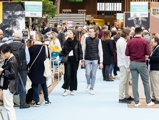 Allée animée d’un salon professionnel avec des visiteurs et exposants échangeant entre des stands de produits gastronomiques, notamment de jambons et de fromages.
