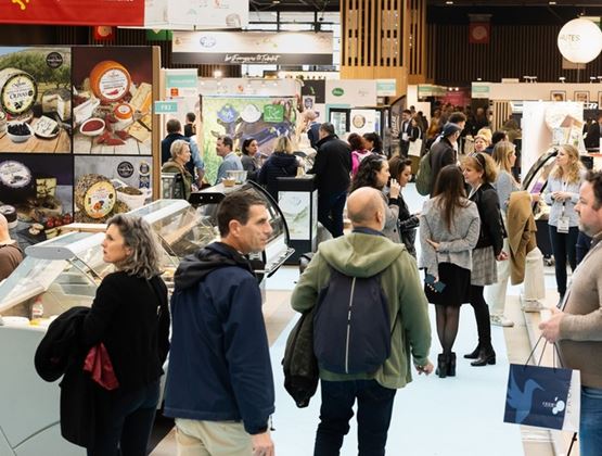 Allée fréquentée d’un salon professionnel alimentaire, avec des visiteurs échangeant devant des stands de fromages et de produits gastronomiques.