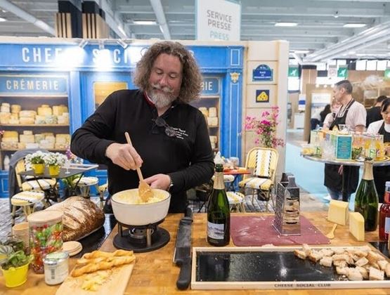 Chef préparant une fondue au fromage sur un stand du Salon du Fromage et des Produits Laitiers, entouré de produits et de visiteurs.