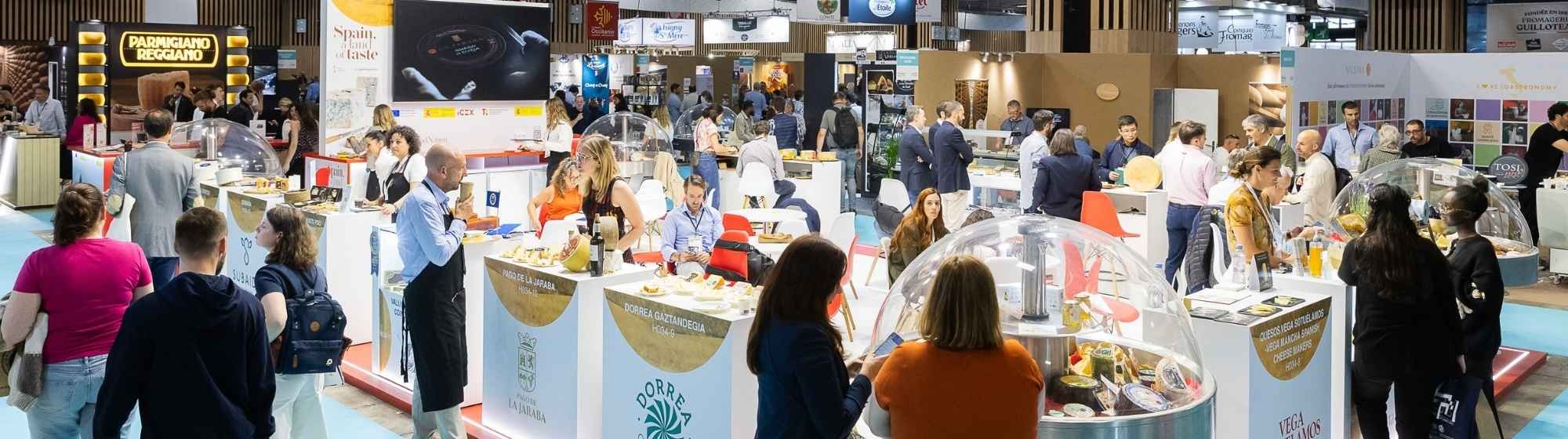 Trade show for cheese and dairy products, featuring numerous exhibitors and visitors interacting around tasting counters. Several booths showcase regional and international cheeses in a vibrant and welcoming atmosphere.