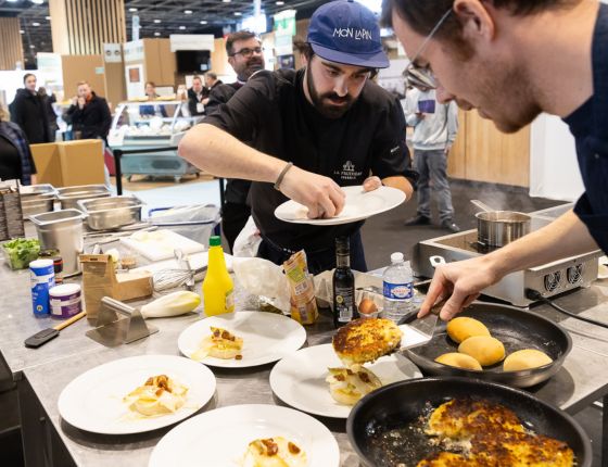 Deux chefs en train de préparer des plats de grilled cheese au concours, dans une cuisine avec des ingrédients visibles.