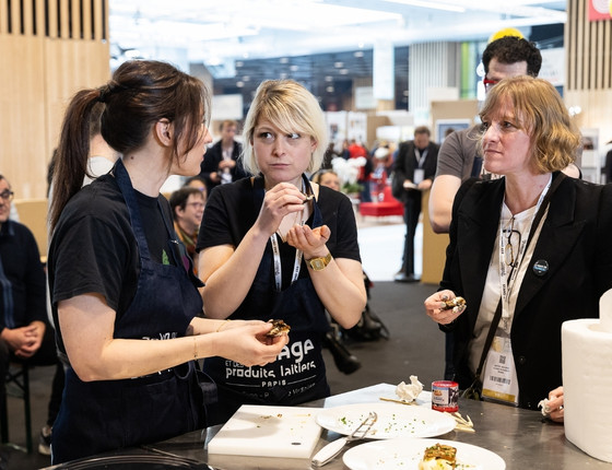 Plusieurs participants en train de cuisiner et de servir des plats lors d'un événement, avec des produits et des ustensiles de cuisine visibles.